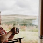 Home Brewing - Side view of young dreamy male with mug of hot beverage looking away against glass wall in countryside house