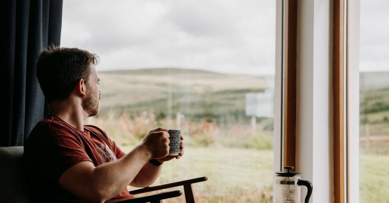 Home Brewing - Side view of young dreamy male with mug of hot beverage looking away against glass wall in countryside house