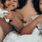 Relationship Challenges - From above of happy African American sisters in domestic wear lying on comfortable bed together and playing video game with gamepads during free time at home