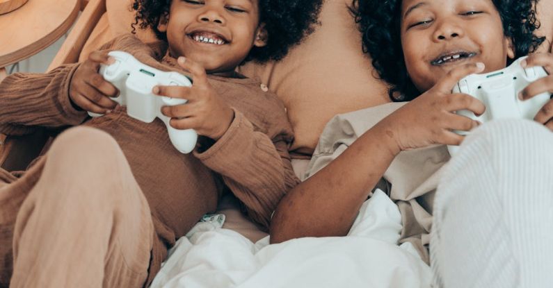 Relationship Challenges - From above of happy African American sisters in domestic wear lying on comfortable bed together and playing video game with gamepads during free time at home