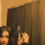 Hair Care Routine - Focused female with dark hair in sweater looking in mirror and curling hair while standing near shower curtain under lamps